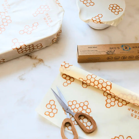 A pair of scissors rests on a yellow honeycomb-patterned beeswax food wrap from Bees Wrap Roll, beside its box and dishes covered with the same wrap, all on a white marble surface.