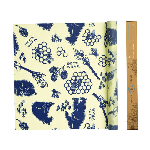 A sheet of Bees Wrap Roll reusable food wrap with a yellow background and blue bears, bees, honeycombs, and berries is placed next to its rectangular brown packaging tube.