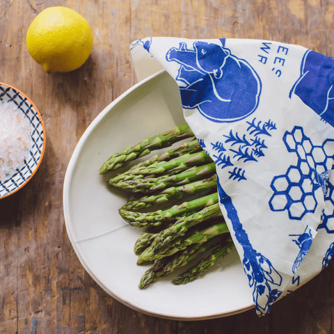 Fresh asparagus spears in a white bowl are partially covered with Bees Wrap Roll, a stylish plastic wrap alternative. A lemon and coarse salt sit nearby on the wooden surface.