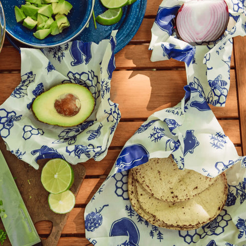 Sliced avocado, red onion, and tortillas are wrapped in Bee’s Wrap Roll—a reusable plastic wrap alternative—on a wooden table with lime wedges and a plate of diced avocado.