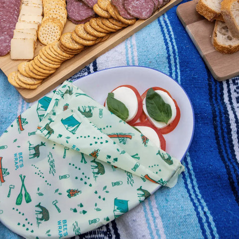 A plate of tomato, mozzarella, and basil is partially covered by Bees Wrap Roll, a reusable beeswax wrap and plastic wrap alternative. Nearby are crackers, cheese, meats on a platter, and sliced bread on a blue patterned blanket.