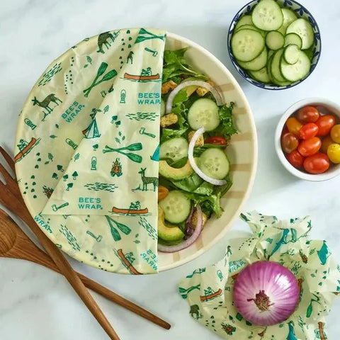 A bowl of salad covered with Bees Wrap Roll—an eco-friendly plastic wrap alternative—beside sliced cucumbers, cherry tomatoes, a wrapped half red onion, and wooden salad utensils on a white surface.