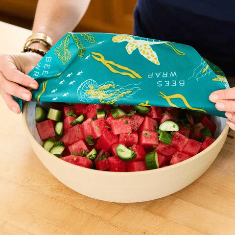 A person covers a bowl of watermelon and cucumber salad with a Bees Wrap Roll—a reusable, eco-friendly alternative to plastic wrap—featuring a teal design with sea creatures and plants.