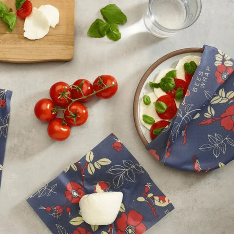 A bowl covered with Bees Wrap Roll, a floral-patterned beeswax wrap by Bees Wrap, sits on a table with mozzarella, basil, and tomatoes. Nearby are a wooden board, glass of water, vine tomatoes, and another wrap holding cheese.