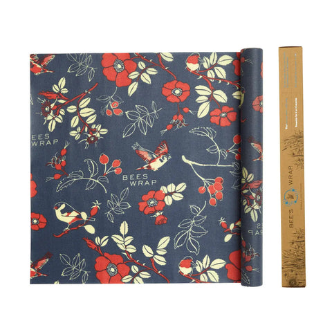 A rolled sheet of Bees Wrap Roll, a reusable food wrap by Bees Wrap, lies on a dark blue background patterned with red flowers, birds, berries, and white leaves. A cardboard tube labeled Bees Wrap is next to it.