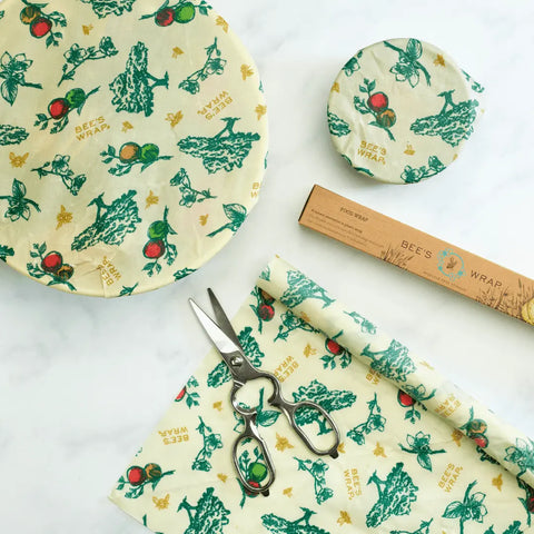 Bees Wrap Roll with green and red fruit patterns covers bowls as a plastic wrap alternative. A pair of scissors, a rolled sheet, and the Bees Wrap Roll box are also shown on a white surface.