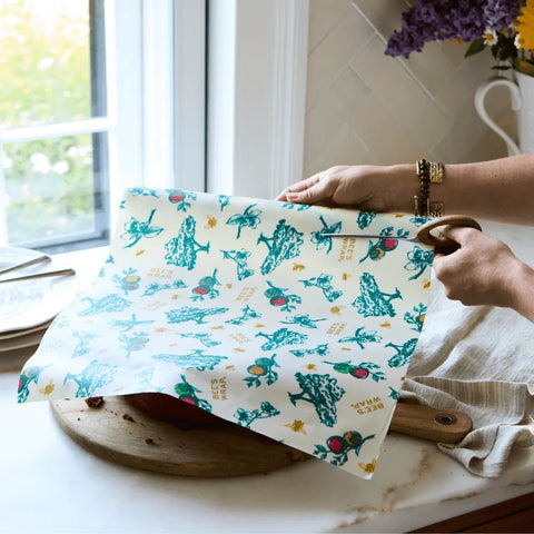 A person covers a bowl with Bees Wrap Roll, a plastic wrap alternative featuring green and yellow vegetable designs, on a kitchen counter near plates, a towel, and a vase of purple flowers by the window.