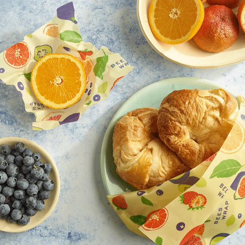 A flat lay shows two croissants on a plate, a halved orange in Bees Wrap Roll (a reusable plastic wrap alternative), blueberries in a bowl, and extra orange halves, all set against a light blue background.