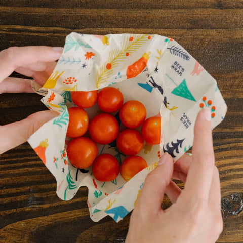 Hands hold Bees Wrap Assorted 3 Pack, a reusable beeswax food wrap by Bees Wrap, filled with small red tomatoes to keep them fresh without plastic on a dark wooden surface.