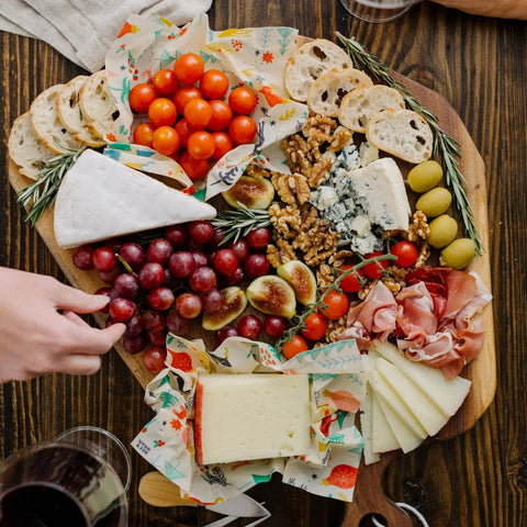 Bee’s Wrap Assorted 3 Pack keeps bread, cheese, fruit, and meats fresh—plastic-free—on a wooden board. Glasses of red wine rest on the table as someone reaches for food.