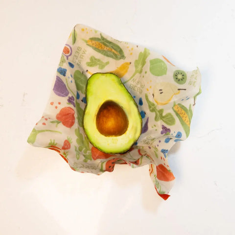 A halved avocado with its pit sits on a Bees Wrap Assorted 3 Pack beeswax wrap, featuring colorful fruit and veggie prints, keeping your food fresh without plastic on a clean white surface.