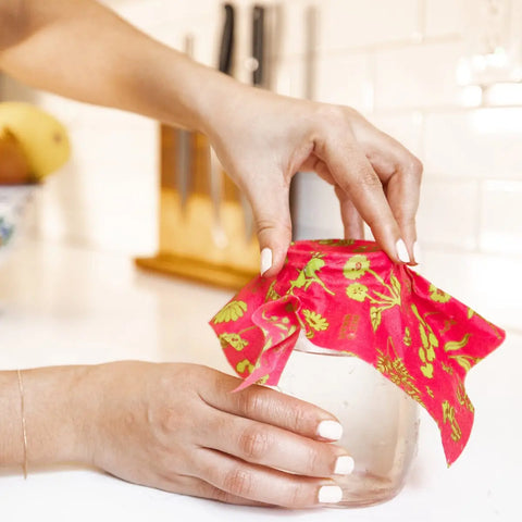 In a modern kitchen, someone uses Bees Wrap Assorted 3 Pack—a plastic-free beeswax food wrap with yellow-green designs—to cover a glass jar and keep food fresh.