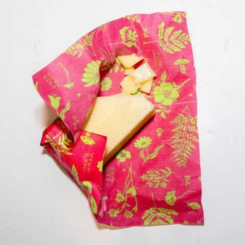 A piece of cheese partially wrapped in Bees Wrap Assorted 3 Pack, a plastic-free pink beeswax food wrap with yellow-green floral patterns, sits on a white surface to keep food fresh.