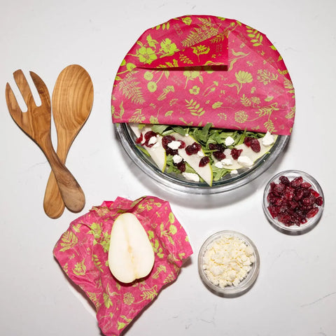 A bowl of salad with pear slices, cheese, and cranberries is partially covered by Bees Wrap Assorted 3 Pack pink floral beeswax wrap to keep it fresh, with wooden servers and small bowls of dried cranberries and cheese nearby.