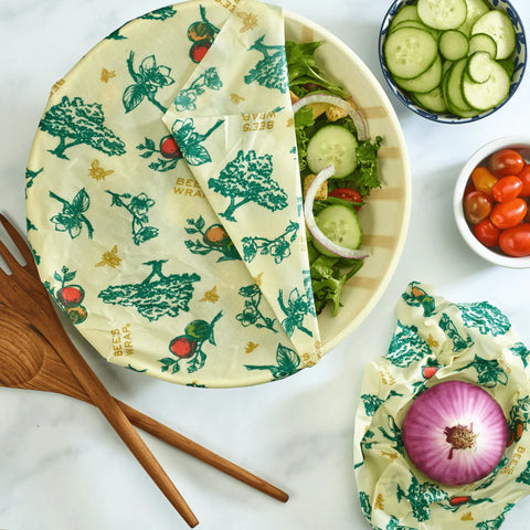 A salad bowl covered with Bees Wrap reusable beeswax wrap from the Assorted 3 Pack sits on a marble countertop, keeping food fresh without plastic. Nearby are sliced cucumbers, cherry tomatoes, and a halved red onion wrapped in Bee’s Wrap.