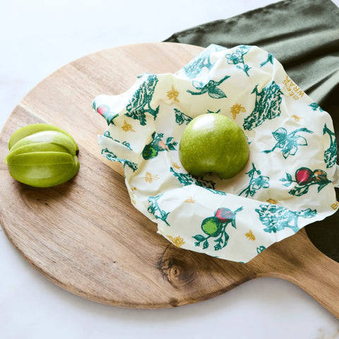 A green apple rests on a floral Bees Wrap from the Assorted 3 Pack, keeping food fresh and plastic-free atop a wooden cutting board. Another green apple sits nearby, next to a dark green napkin.