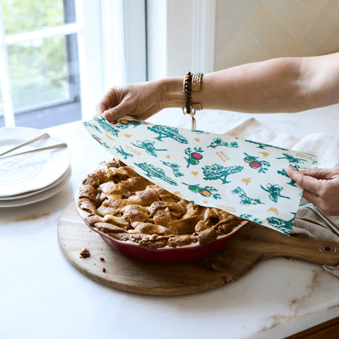 Standing at a kitchen counter near stacked plates and a window, a person uses Bees Wrap Assorted 3 Pack reusable food wrap printed with blue and green illustrations to cover a baked pie in a red dish, keeping food fresh without plastic.