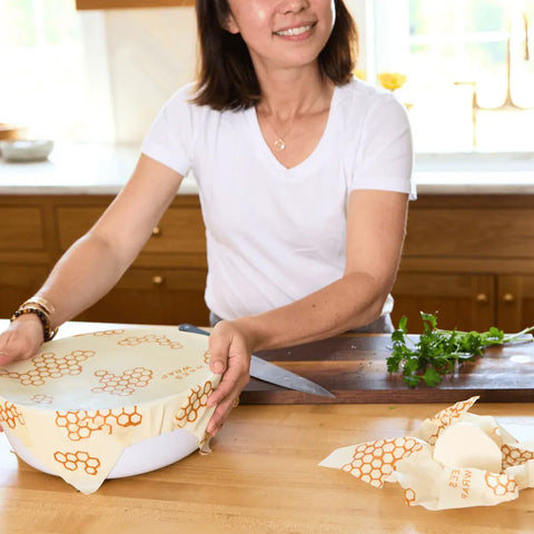 In a bright kitchen, a woman in a white t-shirt covers a bowl with a Bees Wrap Assorted 3 Pack beeswax wrap, keeping food fresh. Fresh cilantro and extra plastic-free Bees Wraps sit on the wooden counter nearby.