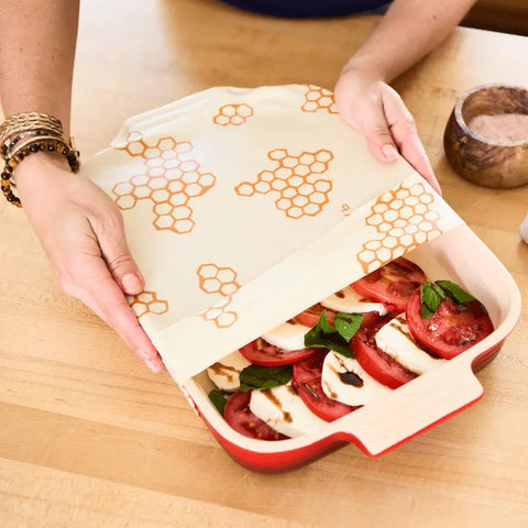 A person covers sliced tomatoes, mozzarella, and basil with a Bees Wrap Assorted 3 Pack beeswax food wrap to keep their dish fresh on a wooden countertop.