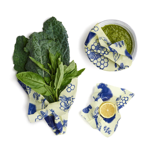 Bees Wrap Assorted 3 Pack features a blue and white animal and flower design on pale yellow, keeping food fresh and plastic-free. Shown wrapping leafy greens, a bowl of green sauce, and a lemon slice.