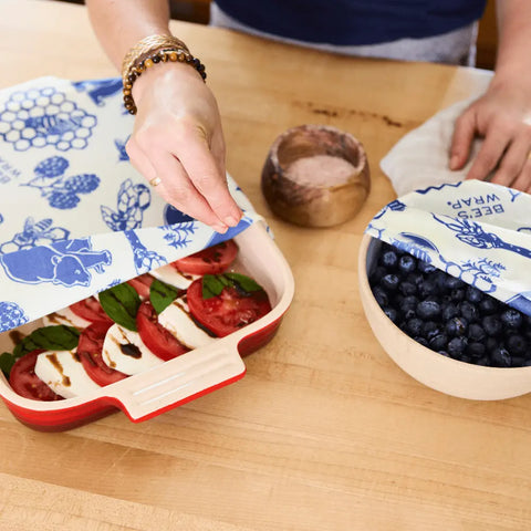 A person uses the Bees Wrap Assorted 3 Pack to keep sliced tomatoes, mozzarella, basil, and balsamic glaze fresh, and also covers a bowl of blueberries on the wooden countertop—all without plastic.