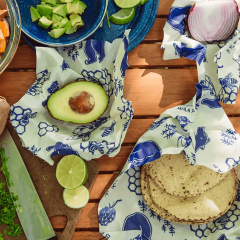 A wooden table with bowls of avocado, lime, cilantro, and red onion. Tortillas and half an avocado stay fresh in Bees Wrap Assorted 3 Pack blue-and-white plastic-free wraps featuring animal and bee designs.