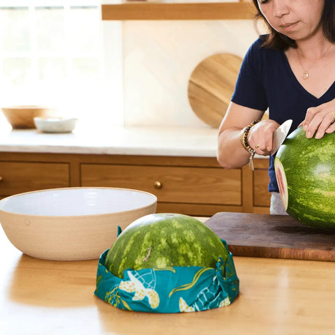 A woman slices a watermelon on a cutting board, while half sits fresh on the counter wrapped in Bee’s Wrap Assorted 3 Pack—an eco-friendly, plastic-free food wrap.