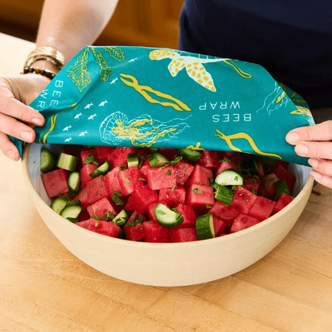 A person covers a bowl of chopped watermelon and cucumber with the Bee’s Wrap Assorted 3 Pack, featuring sea turtle and seaweed designs, to keep food fresh without plastic.