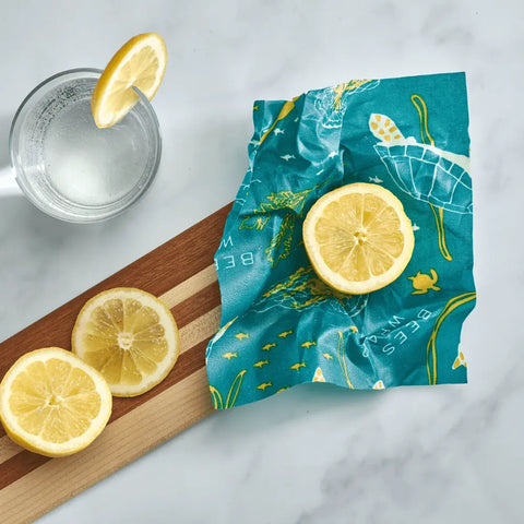 A glass of water with lemon sits beside a wooden board holding lemon slices and a Bee’s Wrap Assorted 3 Pack reusable, plastic-free wrap by Bee’s Wrap with sea turtle designs, keeping food fresh on a marble surface.