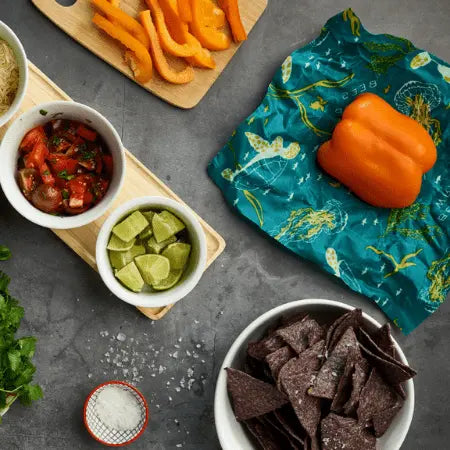 A top-down view of a tabletop with diced tomatoes, lime wedges, herbs, salt, blue corn tortilla chips, and orange bell peppers on a Bees Wrap Assorted 3 Pack plastic-free beeswax wrap to keep food fresh.