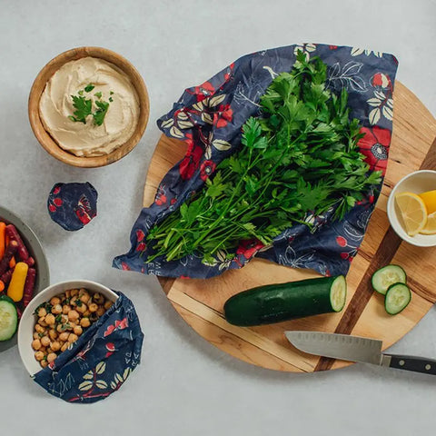 Fresh parsley and a cucumber sit on a wooden cutting board with a knife, near bowls of hummus, salad, and lemon wedges. The food is partially covered with Bees Wrap Assorted 3 Pack plastic-free floral wraps.