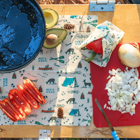 Chopped onions and sliced red pepper on a cutting board sit beside an avocado, pinecone, and Bees Wrap Assorted 3 Pack food wraps—reusable, camping-themed wraps perfect for keeping food fresh and plastic-free on a wooden table.