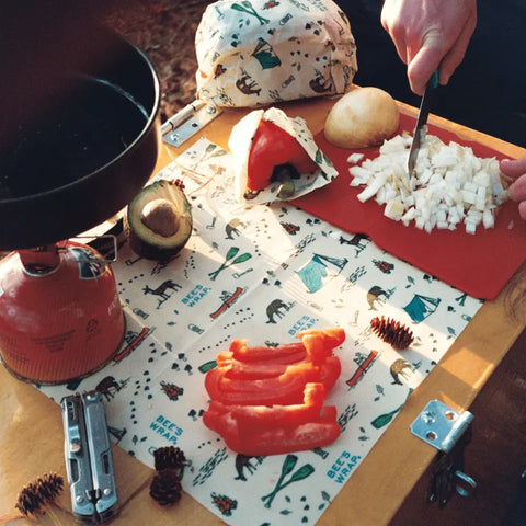 A person chops onions next to sliced red bell peppers, an avocado, a camping stove, pinecones, a multitool, and Bees Wrap Assorted 3 Pack plastic-free containers on a table with a colorful nature-themed cloth.
