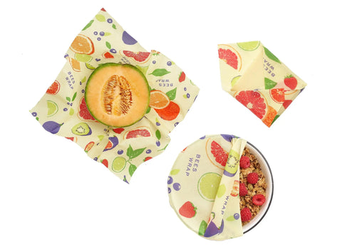 Three pieces from the Bees Wrap Assorted 3 Pack cover a melon half, granola with raspberries, and a folded wrap, keeping food fresh plastic-free. The wraps feature fruit patterns and BEES WRAP on a white background.