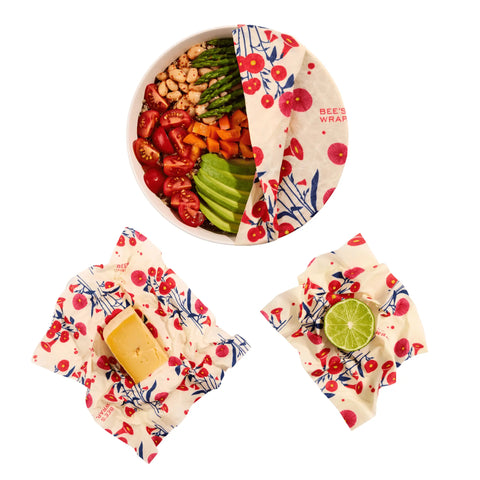 A salad bowl, cheese block, and halved lime stay fresh under Bees Wrap Assorted 3 Pack plastic-free wraps with a red and blue floral pattern by Bees Wrap, displayed on a white background.