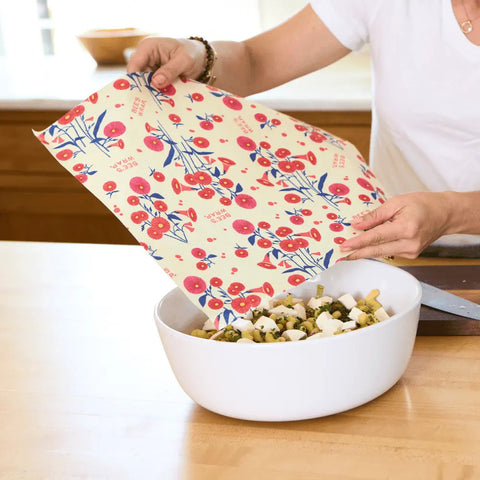 In a kitchen, someone covers a bowl of chopped salad with Bees Wrap Assorted 3 Pack—reusable, floral-patterned wraps that keep food fresh without plastic.