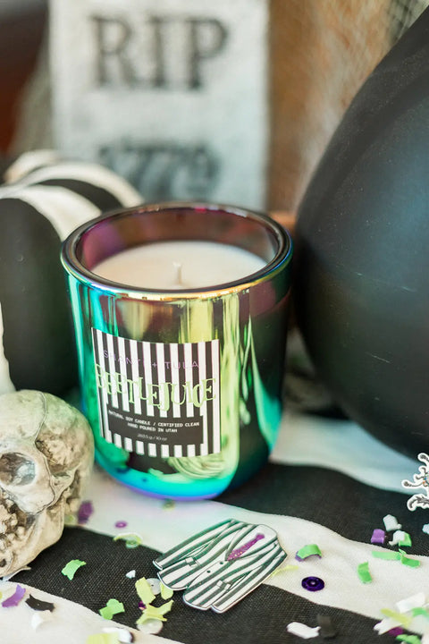 A Shanti + Tula Beetlejuice Halloween Candle—White Birch, Cranberry & Orange Peel, hand-poured soy wax and made in the USA—sits among Halloween decor like a skull, confetti, a striped coffin pin, and a RIP tombstone sign.