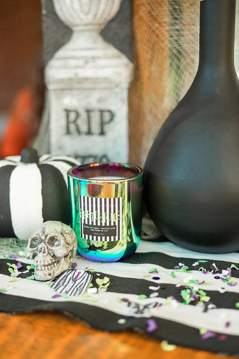 A Halloween scene features a shiny green hand-poured candle, small skull, black vase, and Shanti + Tula’s Beetlejuice Halloween Candle (White Birch, Cranberry & Orange Peel), plus an RIP gravestone prop and confetti on a striped cloth.