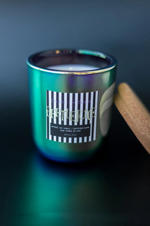 The Shanti + Tula Beetlejuice Halloween Candle features hand-poured white soy wax in a metallic green and purple jar labeled “Beetlejuice” on black-and-white stripes, with a cork lid beside it. Scent: White Birch, Cranberry & Orange Peel. Made in USA.