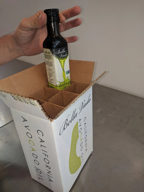 A hand places a bottle of Bella Vado Avocado Oil into a Case of Conventional Oil (6 Bottles) - 10% Discount, marked CALIFORNIA AVOCADO OIL, resting on a metal surface.