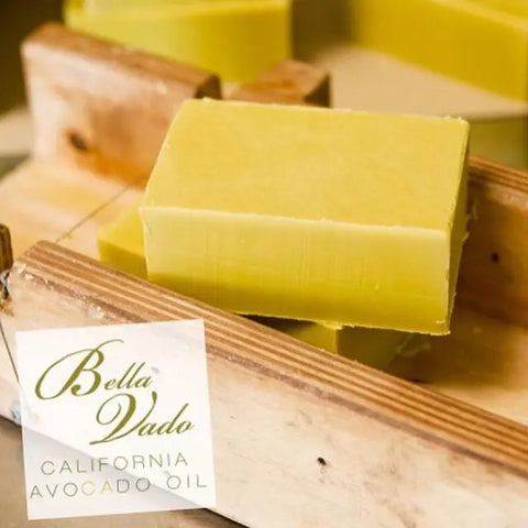 Moisturizing yellow-green bars of Bulk Soap by the Case - Select 6, made with Bella Vado Avocado Oil, rest on a wooden soap cutter. The white label features Bella Vado Avocado Oil in elegant script.
