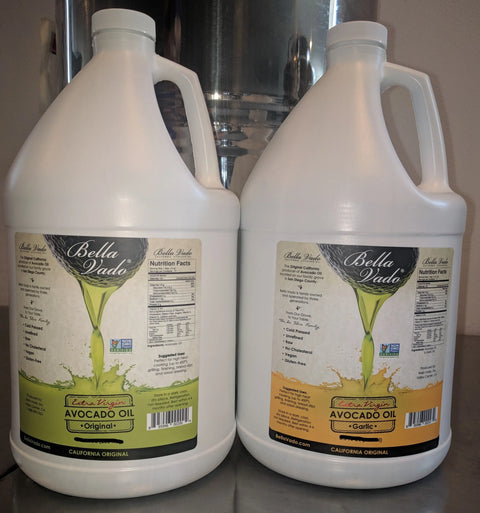 Two gallon jugs of Bella Vado Avocado Oil Extra Virgin—Original (green label) and Garlic (yellow label)—are placed on a metal surface.