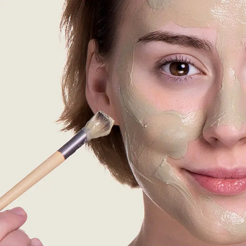 A woman with short brown hair applies Open Formula Bentonite Clay Mask to her face with a brush, smiling slightly. Only half of her face is visible against a light background. This mask exfoliates and reduces acne scars.