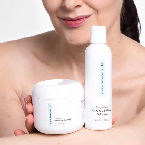 A woman with bare shoulders smiles slightly while holding two skincare products: Open Formula’s Bentonite Clay Mask, famed for reducing acne scars, and a white bottle labeled AHA Mud Mask Solution.