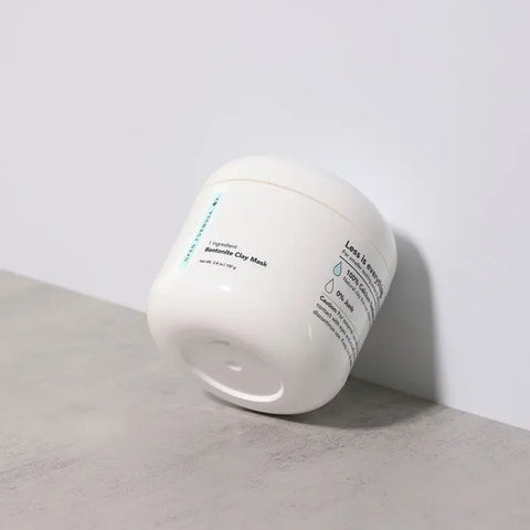 A white jar of Open Formula Bentonite Clay Mask, containing exfoliating bentonite clay to help reduce acne scars, rests on its side against a white wall and gray surface with ingredient and usage info visible on the label.