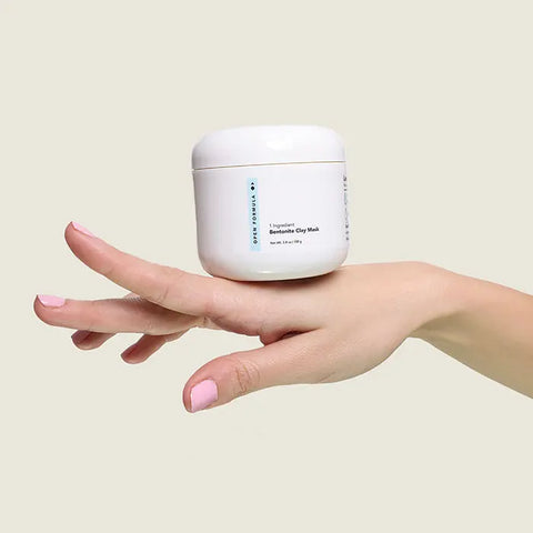 A hand with light pink nail polish balances a white jar of Open Formula Bentonite Clay Mask on its fingers against a plain beige background.