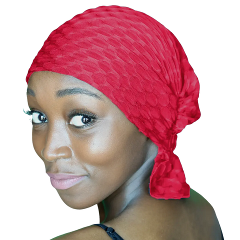 A woman with glowing brown skin smiles gently over her shoulder, wearing the Chemo Beanies® Kendall textured red chemo beanie tied at the back. Her subtle makeup and highlighted eyes stand out against a black background.