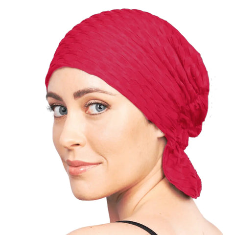 A light-skinned, blue-eyed woman wears the bright red Kendall Chemo Beanie® by Chemo Beanies®, tied at the back. She looks over her shoulder and smiles slightly against a white background.