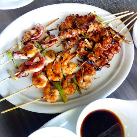 A white plate with skewers of grilled shrimp, squid, and glazed meat seasoned with Gustus Vitaes Better Than Butter Seasoning Set, served with a small bowl of soy sauce on the side.