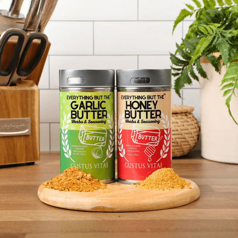 Two tins from the Gustus Vitae Better Than Butter Seasoning Set (2 Pack) sit on a kitchen counter, surrounded by kitchen items and a plant, with piles of seasoning displayed in front.